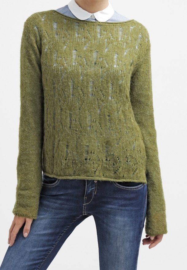 Personne portant un pull vert tricoté à manches longues par-dessus une chemise bleue à col et un jean bleu, debout avec une main détendue.