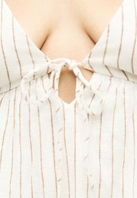 Λευκό ύφασμα με λεπτές κάθετες μπεζ ρίγες δεμένο σε φιόγκο πάνω σε λαιμόκοψη με άνοιγμα σε ανοιχτόχρωμο δέρμα.