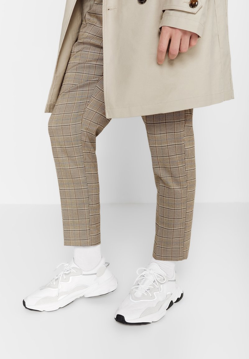 Gabardina beige sobre pantalones de cuadros marrones con un corte ajustado. Lleva zapatillas blancas con paneles texturizados y suela negra.