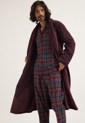 Un homme barbu porte un long peignoir bordeaux sur un pyjama assorti à carreaux bleus et bordeaux, devant un fond de studio uni.