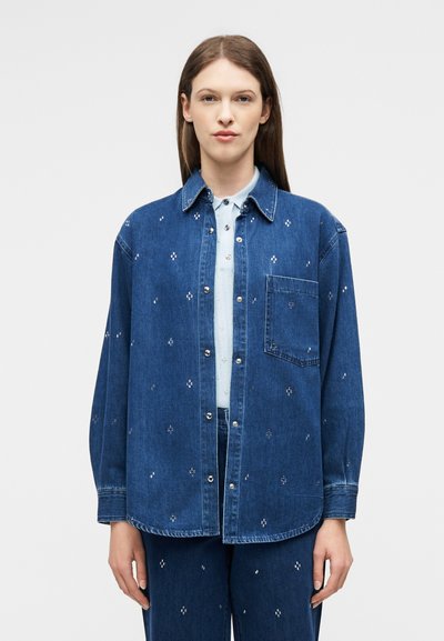 Femme portant une veste en jean bleue et un pantalon avec de petits motifs floraux blancs, debout devant un fond clair uni.