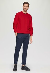 Rode sweatshirt met ronde hals en omgeknoopte mouwen, gecombineerd met marineblauwe chinos. Zwarte sneakers maken de look compleet. Eenvoudige, casual outfit.