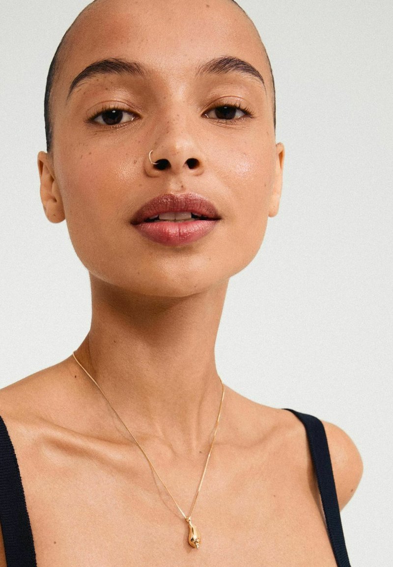Collar de oro con un colgante en forma de lágrima sobre una delicada cadena. La modelo tiene la piel suave y un fondo neutro.