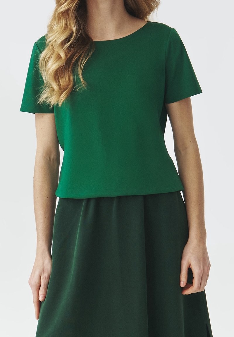 Femme portant un haut vert à manches courtes rentré dans une jupe vert foncé, avec des cheveux blonds ondulés sur l'épaule.