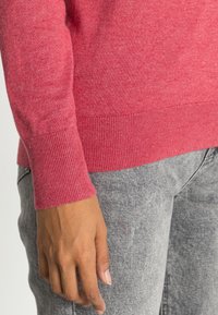 Main reposant sur le côté, portant un pull rose à manches longues et un jean en denim gris clair.