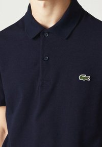 Mörkblå polo skjorta i texturerat tyg med klassisk krage, som har två knappar och en broderad grön krokodil-logotyp på bröstet.