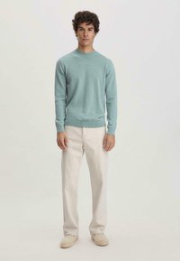 Hellblaues Strickpullover mit Rundhalsausschnitt, kombiniert mit cremefarbenen Hosen. Das Modell steht vor einem schlichten Hintergrund.