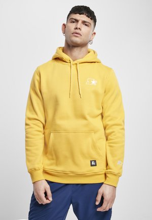 Žltá mikina s veľkou prednou vreckom, kapucňou so šnúrkou a logom na hrudi. Má rebrované manžety a lem. Vyrobená z mäkkej bavlnenej zmesi.