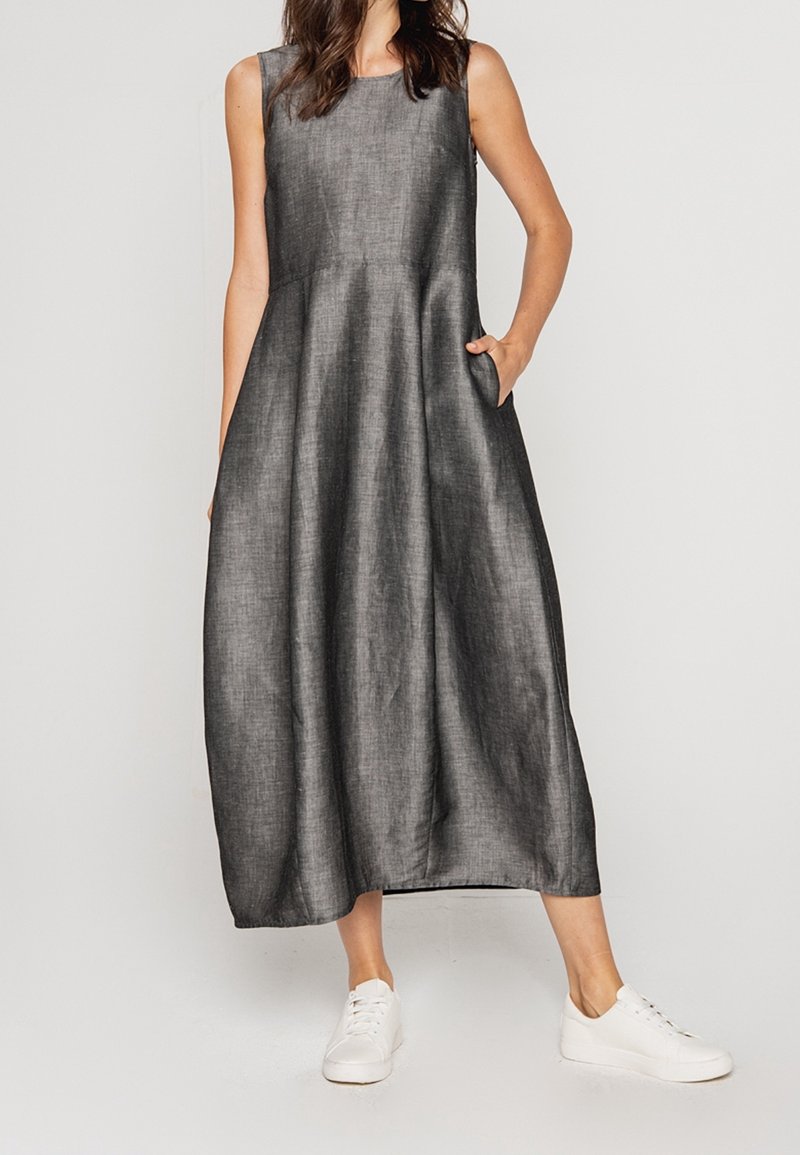Femme portant une robe longue sans manches en lin gris foncé avec des poches et des baskets blanches, debout devant un fond uni.