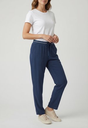 Donna con outfit casual che indossa una t-shirt bianca a maniche corte, pantaloni con coulisse blu navy e sneakers beige, in piedi contro uno sfondo semplice.