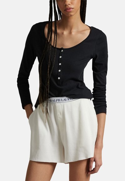 Femme portant un haut henley noir à manches longues et un short blanc côtelé avec une ceinture Polo Ralph Lauren visible, dans une pose décontractée.