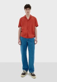 Rotes Kurzarm-Hemd mit Knopfleiste und Kragen, kombiniert mit hellblauen, geraden Jeans und braunen Sneakern mit weißen Sohlen.