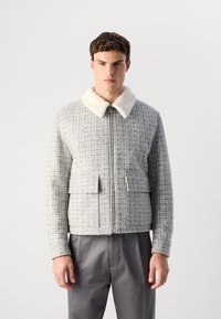 Bărbat îmbrăcat cu o jachetă gri cu carouri și fermoar, cu guler din fleece alb și buzunare mari în față, purtând pantaloni gri, stând cu spatele la un fundal alb.