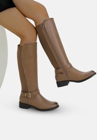 Piernas con botas marrón claro hasta la rodilla, con cremallera lateral y detalle de hebilla, un pie levantado, fondo claro y sencillo.
