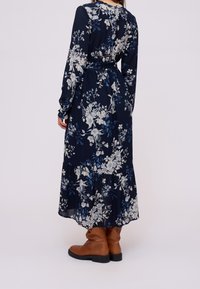 Robe longue bleu marine à motif floral crème et bleu. Présente des manches longues, un ourlet à volants et une taille cintrée. Portée avec des bottes marron montantes jusqu'aux genoux.