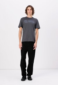 Camiseta de algodón gris con mangas cortas y un escote redondeado, con texto azul impreso. Combinada con pantalones negros y zapatos negros.
