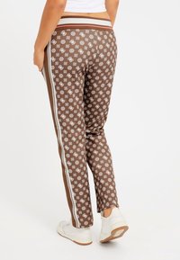 Brown patterned joggers with white accents and side stripes, featuring a high waistband and a slim fit. Worn with white sneakers.