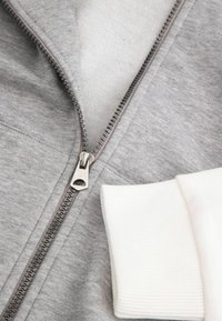 Szary sweatshirt zapinany na zamek błyskawiczny z metalowym suwakem, teksturowanym materiałem i białymi ściągaczami. Zbliżenie na detal zamka i ściągacza.