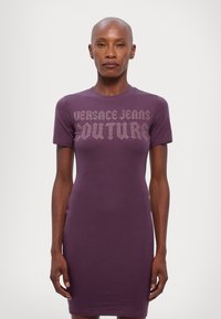 Fialové upnuté šaty ve tvaru trička s krátkým rukávem. Na přední straně se nachází text "VERSACE JEANS COUTURE" v kontrastním designu. Hladká textura látky.