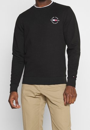 Muž v černé mikině Tommy Hilfiger s dlouhým rukávem a logem na hrudi a rukávu, v béžových kalhotách, stojící před bílým pozadím.