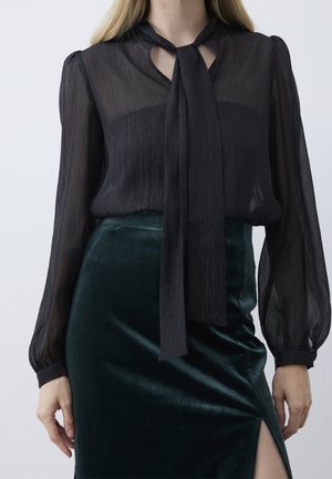 Femme portant un chemisier noir transparent à manches longues avec un nœud papillon noué au col et une jupe en velours vert foncé fendue sur la cuisse.