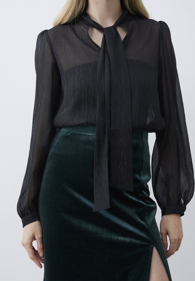 Femme portant un chemisier noir transparent à manches longues avec un nœud papillon noué au col et une jupe en velours vert foncé fendue sur la cuisse.
