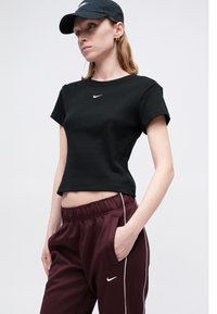 Crni rebrasti skraćeni majici s bijelim Nike logotipom, upareni s burgundskim hlačama s elastičnim pojasom koje imaju bijele bočne trake i džepove.