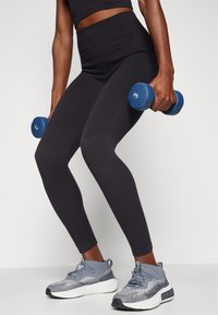 Leggings neri a coste, sneakers grigie con motivi e accenti neri, e manubri blu contrassegnati con il numero "3" tenuti in mano.