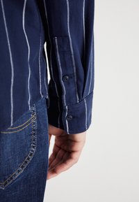 Μπλούζα με μακριά μανίκια σε ναυτικό μπλε χρώμα με ρίγες, με κουμπωτό μανσέτα και υφή υφάσματος. Φορεμένη με τζιν παντελόνι που έχει κίτρινες ραφές στην τσέπη.