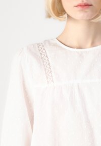 Bílá bavlněná blůza s texturovanými puntíky, krajkovými detaily na rameni a kulatým výstřihem se nařaseným materiálem.