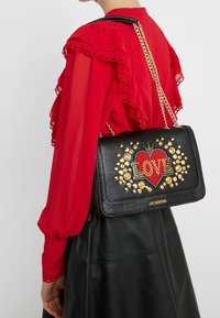 Person som bär en röd volangblus och svart läderskjorta, bär en svart Love Moschino-väska med gyllene nitar och röd hjärtdesign på axelremmen.