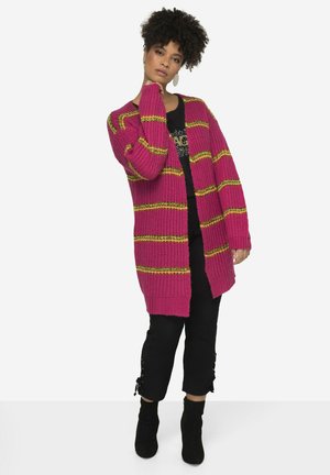 Magenta Strickjacke mit gelben und grünen Streifen, offene Vorderseite, gerippte Textur, kombiniert mit einem schwarzen Oberteil und schwarzen cropped Hosen.