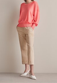 Kobieta w koralowej bluzce z długim rękawem, beżowych spodniach 3/4 i białych sandałach na obcasie, stojąca przy gładkiej beżowej ścianie.