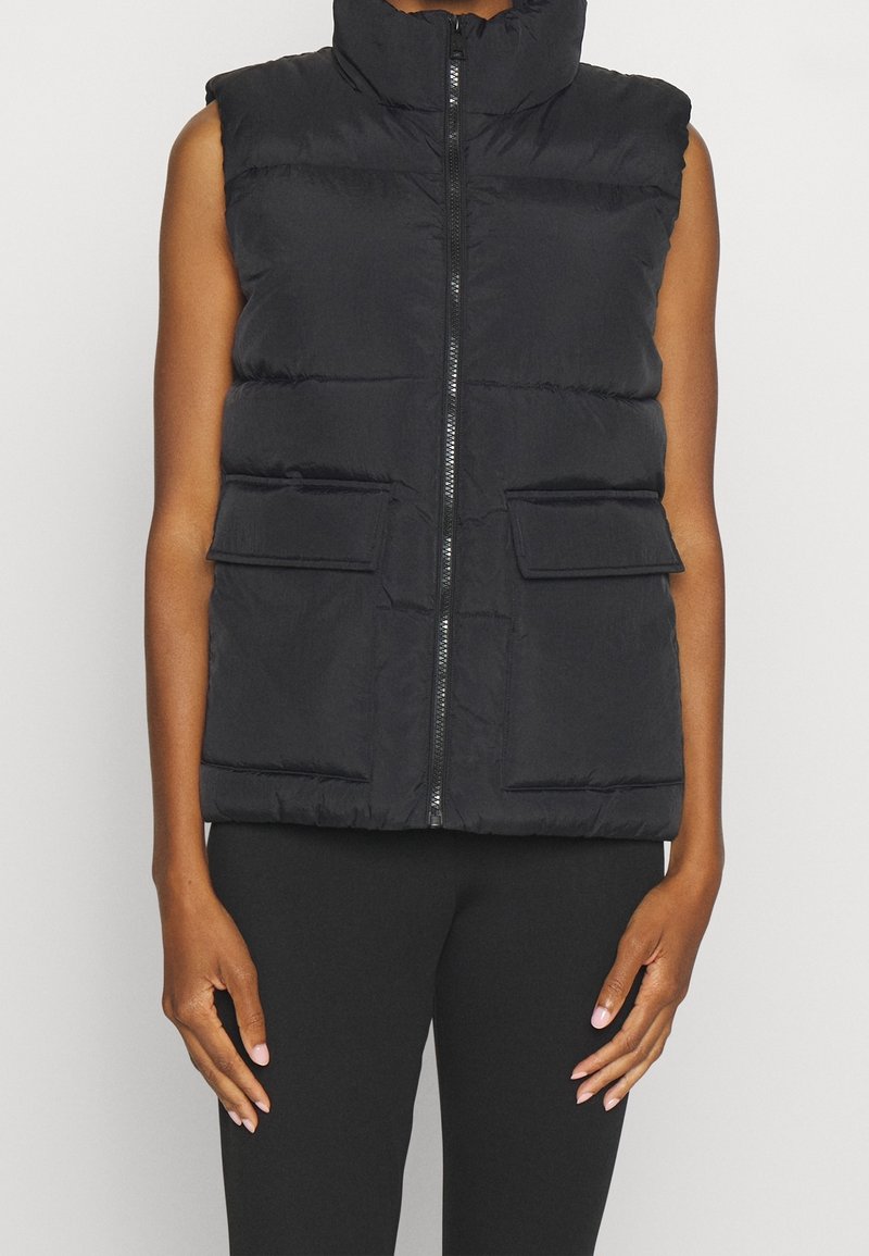 Smanicato nero imbottito con collo alto, zip frontale e due tasche anteriori. Design trapuntato con texture liscia. Stile senza maniche.