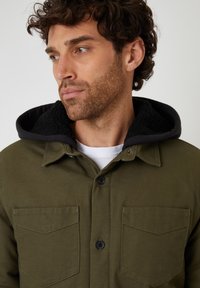 Olivgrön skjortjacka med en svart fleecefodrad huva, som har två bröstfickor och svarta knappar. Tyget ser robust och strukturerat ut.