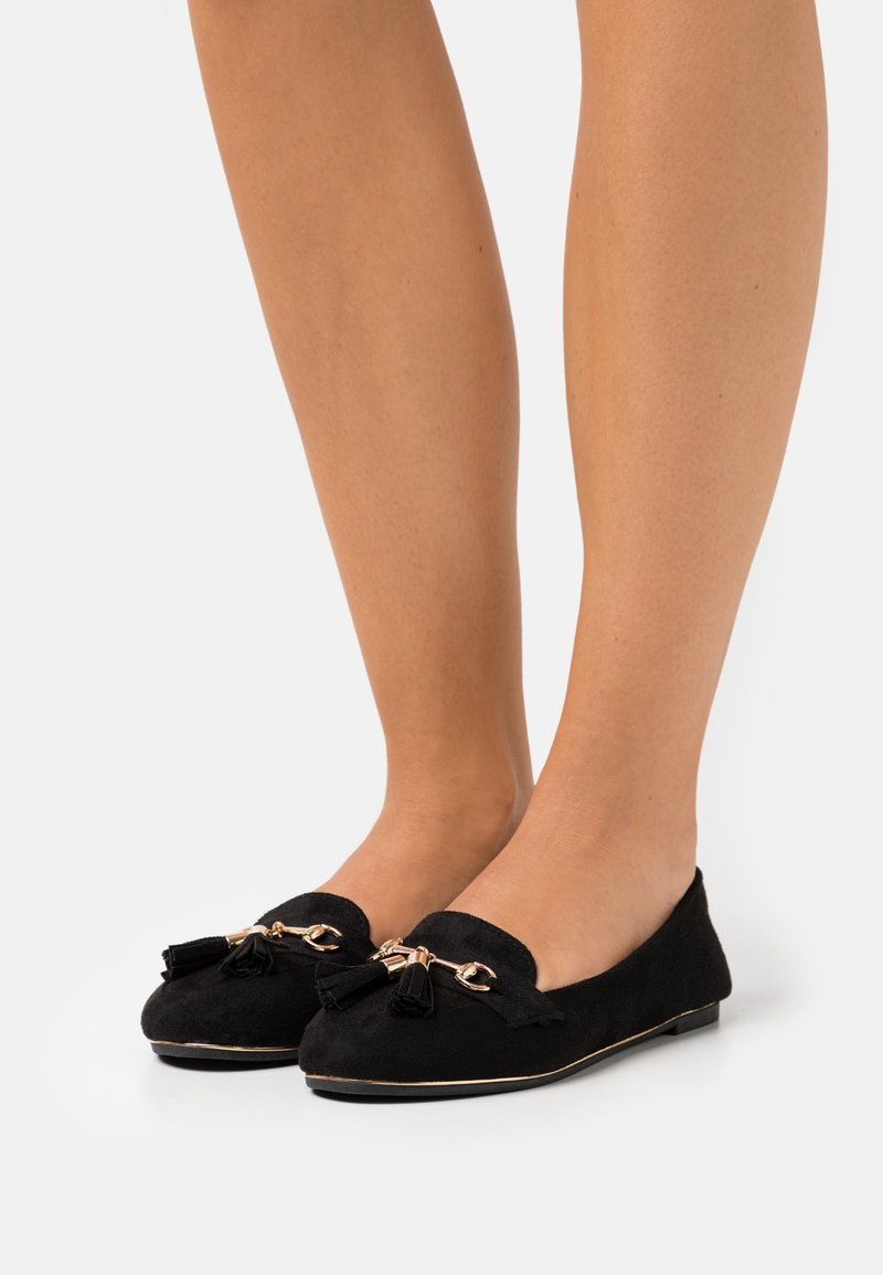 Mocassins en daim noir avec chaîne dorée et détails à pompons portés pieds nus sur fond blanc.
