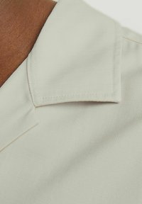 Collared shirt in light beige fabric with a textured ribbed pattern. Close-up view highlighting collar shape and stitching details.