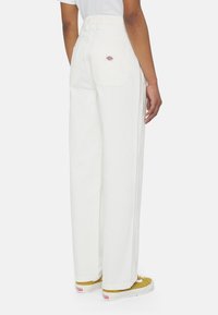 Personne portant un pantalon blanc taille haute à jambes larges avec poches arrière et écusson logo, associé à une chemise blanche et des baskets jaunes.