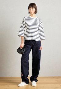 Ριγέ, μακρυμάνικη βαμβακερή μπλούζα με μεγάλο λαιμό, συνδυασμένη με σκούρες τζιν παντελόνες με λουλουδάτα κεντήματα και λευκά αθλητικά παπούτσια. Μαύρη τσάντα χειρός στο χέρι.