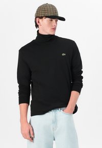 Un pull col roulé noir à manches longues, avec un petit logo vert, associé à un jean bleu clair et une casquette à carreaux marron.
