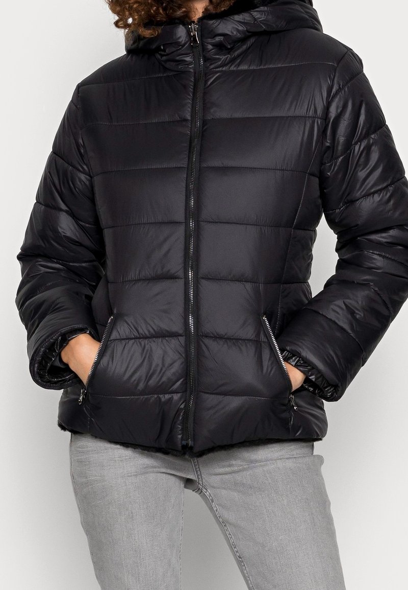 Personne portant une doudoune matelassée noire avec capuche et les mains dans les poches avant zippées, associée à un jean gris clair.