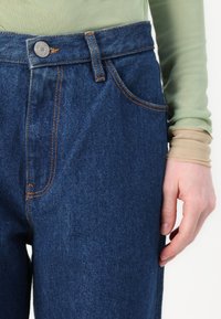 Srednje visoki modri traper hlače z oranžnimi šivi in enim sprednjim žepom. Imajo kovinsko zaponko in ravno obliko hlačnic.