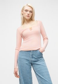 Νεαρή γυναίκα με μακριά ξανθά μαλλιά, που φοράει ροζ μακρυμάνικη μπλούζα με λαιμόκοψη σε σχήμα V και φαρδιά τζιν παντελόνια με μπλε σχέδια.