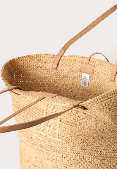 Sac cabas en paille tissée beige avec des poignées en cuir couleur naturel et une étiquette à l'intérieur indiquant les instructions de soin.