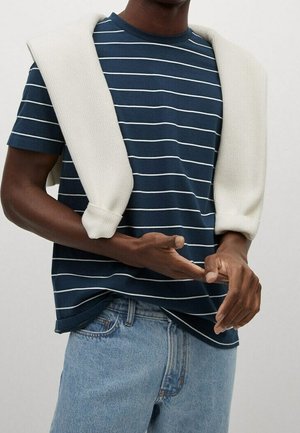 Granatowa koszulka w paski z białymi liniami, połączona z kremowym, dzianym swetrem narzuconym na ramiona, zestawiona z jasnoniebieskimi dżinsami.