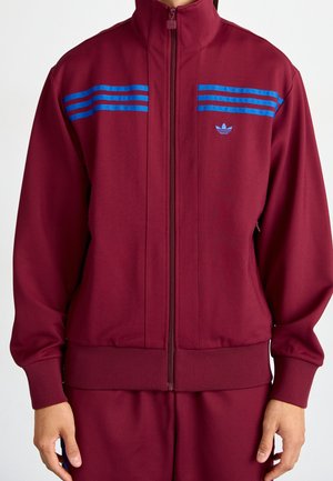 Veste de survêtement bordeaux à fermeture éclair avec trois bandes bleues sur la poitrine et petit logo trèfle sur le côté gauche, portée par une personne.