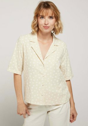 Femme aux cheveux blonds mi-longs portant une chemise crème à boutons avec de petits motifs floraux blancs et un pantalon de couleur claire.