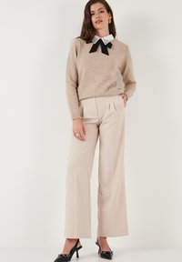 Donna che indossa un maglione beige con colletto bianco decorato e fiocco nero, pantaloni larghi crema e tacchi neri, in piedi contro un muro bianco.