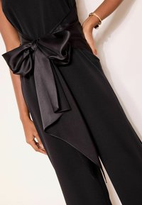 Tuta nera con una cintura larga in satin e un grande fiocco in vita. Il tessuto è liscio e fluente con un design a gamba svasata.