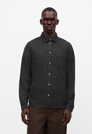Homme portant une chemise en jean gris foncé boutonnée avec une poche poitrine et un pantalon marron sur un fond blanc uni.